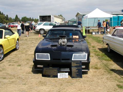 1967 Ford Ranchero - A heavily modified black car with a prominent supercharger is displayed at an outdoor event, alongside other vehicles.