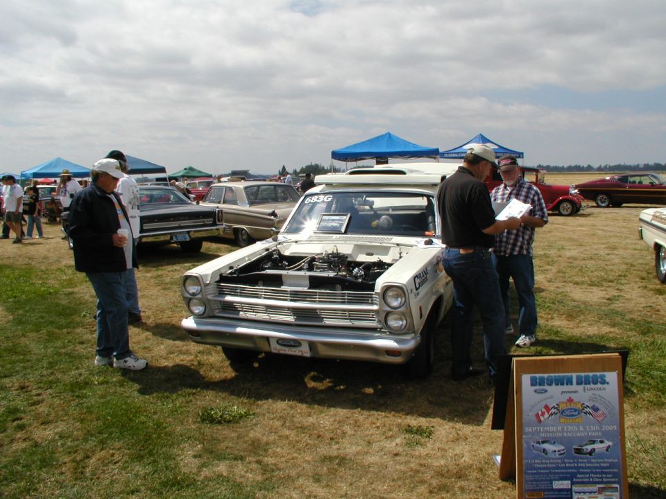 White classic car with hood open, people looking at it. Sign in foreground