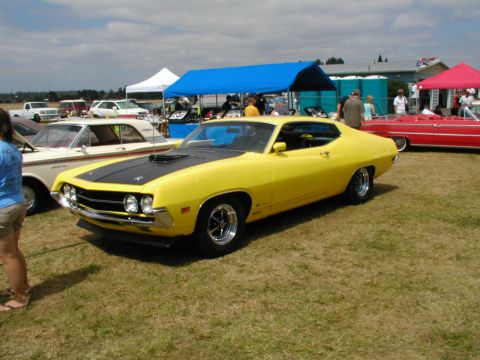 1967 Ford Ranchero - A bright yellow muscle car with a black hood scoop is displayed at a car show, with other vehicles and tents in the background.
