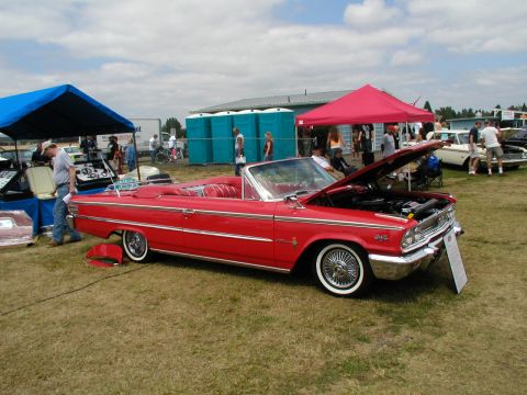 1967 Ford Ranchero - A red convertible with its hood up is displayed at an outdoor car show with other vehicles and tents in the background.