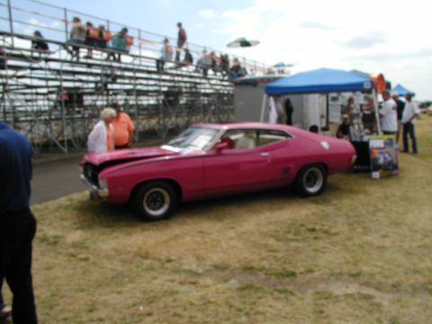 1967 Ford Ranchero - A pink coupe is parked on a grassy field with spectators in bleachers and tents in the background.