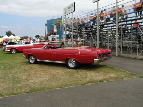 1967 Ford Ranchero - A red convertible is parked on the grass with spectators in the bleachers in the background.