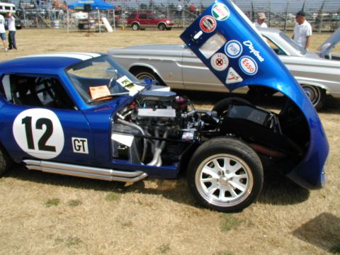 1967 Ford Ranchero - A blue race car with its hood open, displaying the engine and various stickers, sits on a grassy field with spectators in the background.