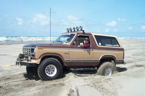 1989 Ford F-150 - This two-tone Ford Bronco is stuck in the sand on a beach, with the ocean in the background.