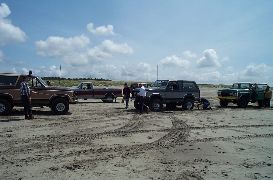 Four vintage Ford Broncos and a pickup parked on beach sand with people gathered around