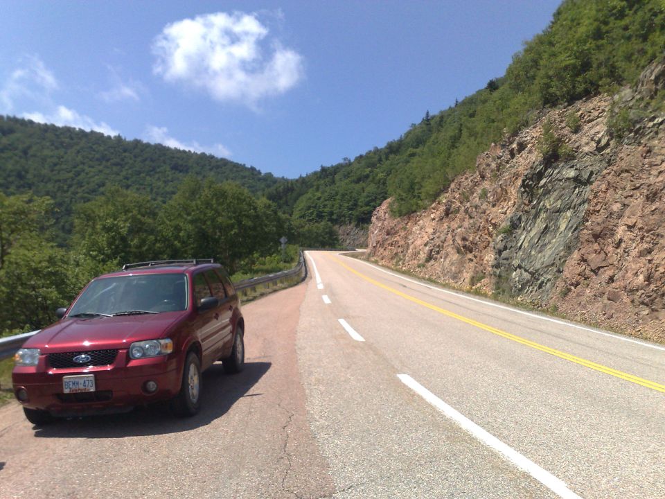 Red 2006 Ford Escape parked on a paved road with a rocky mountainside and green trees