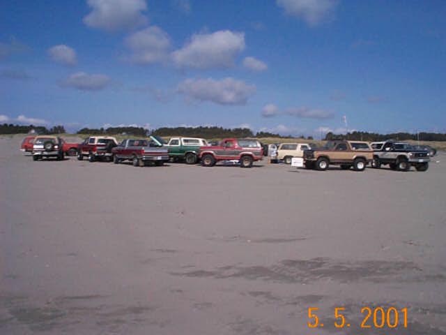 Row of about a dozen lifted 4x4 trucks and Broncos parked on a wide sand beach
