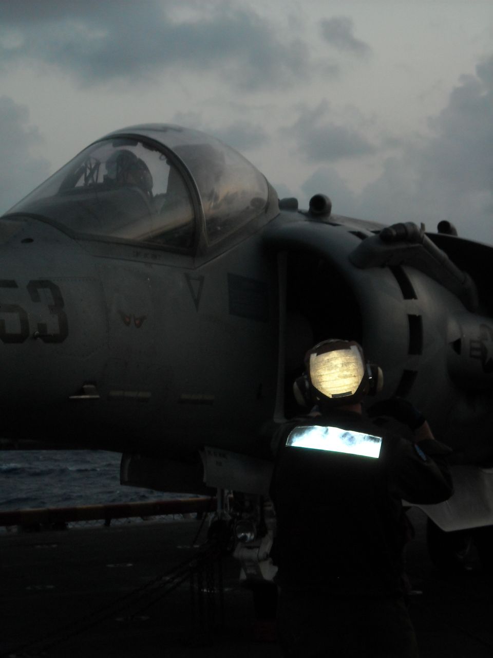 Gray Harrier jet marked '53' on carrier deck with crew member at dusk