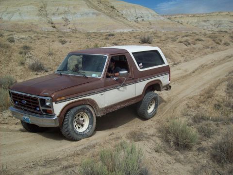1985 Ford Bronco - A two-tone Ford Bronco navigates a dusty trail through a dry, hilly terrain under a partly cloudy sky.