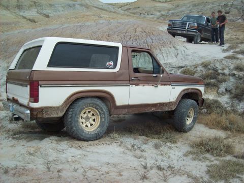 1985 Ford Bronco - A two-tone Ford Bronco navigates a dusty, off-road landscape with another truck visible in the background.