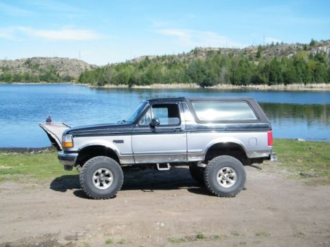 1995 Ford Bronco - A two-tone Ford Bronco is parked on a dirt path next to a body of water with trees and hills in the background.