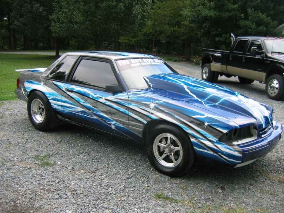 Fox-body Mustang coupe with blue lightning airbrush paint, drag wheels and cowl hood, parked on gravel