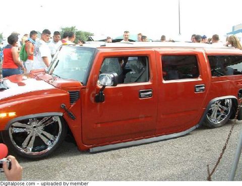 1990 Ford Bronco - Driver-side view of an orange Hummer H2 laid flat on the ground on air suspension at a crowded outdoor show, riding on oversized chrome mul…