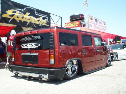 1990 Ford Bronco - Driver-side 3/4 rear view of a candy-red Hummer H2 laid out on air suspension over large chrome wheels at an outdoor car show. The rear gla…