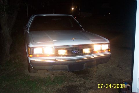1991 Ford Crown Victoria LTD - The front of a white 1991 Ford Crown Victoria LTD is visible at night, with its headlights illuminated.