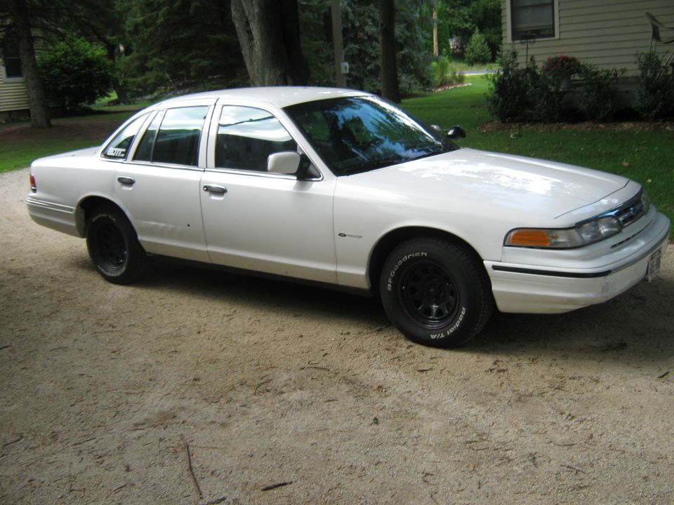 White Crown Victoria sedan front 3/4 view with black steel wheels on gravel driveway