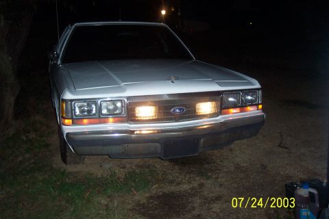 1991 Ford Crown Victoria LTD - The front of a white 1991 Ford Crown Victoria LTD is shown at night with its headlights illuminated.