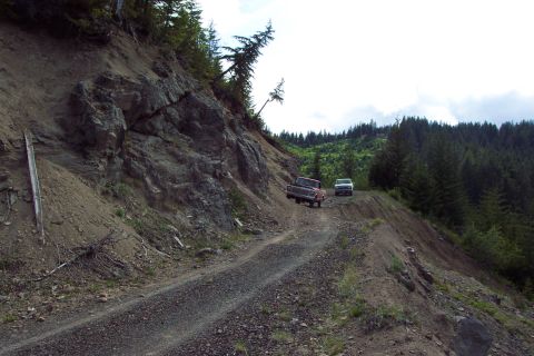 1989 Ford F-150 - Two trucks navigate a precarious dirt road on a steep mountainside with dense forest in the background.