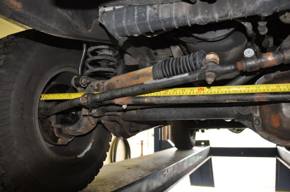 Undercarriage of a vehicle showing steering linkage and a yellow tape measure. Rusty components are visible