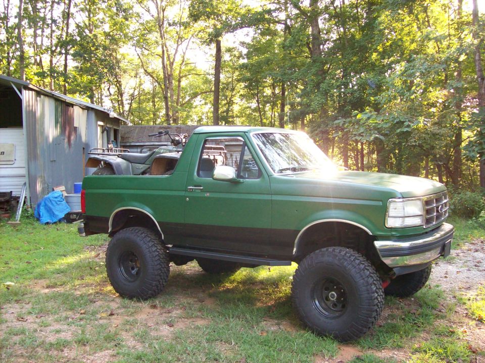 Lifted green topless Ford Bronco, passenger side profile in a wooded yard
