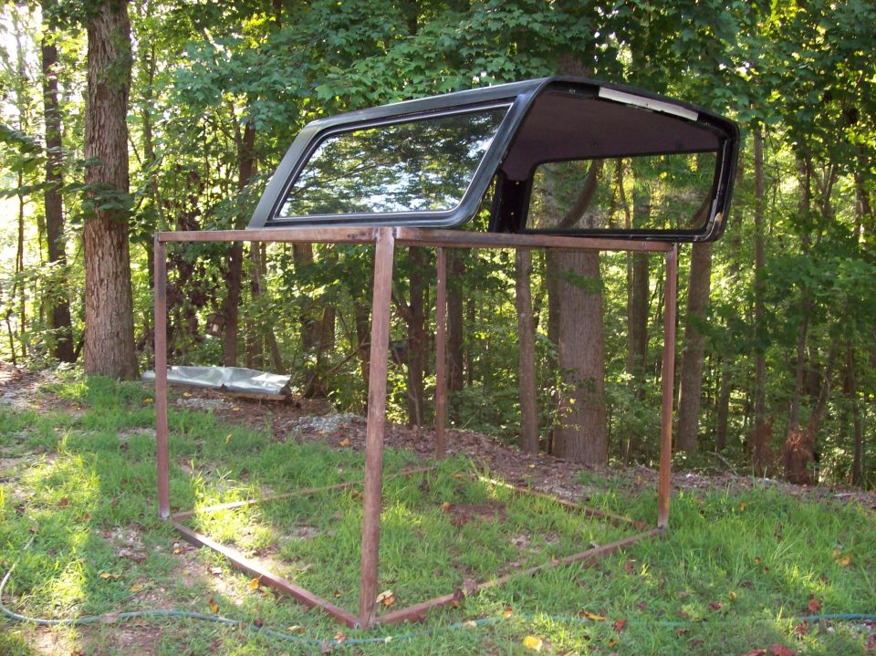 Black Bronco removable hardtop stored on a rusty steel rack in a wooded yard