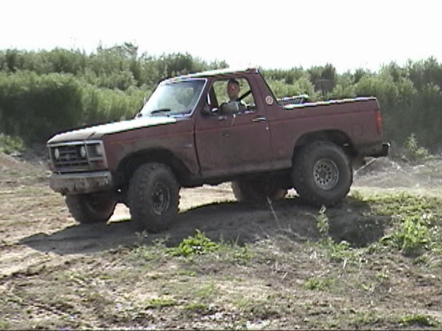 Faded red topless doorless Ford Bronco driven across a rutted dirt clearing