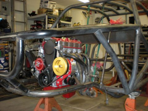 Suzuki Samurai - Bare tube-frame buggy chassis on jack stands in a home shop, with a small red-and-aluminum four-cylinder engine mounted low between the rai…