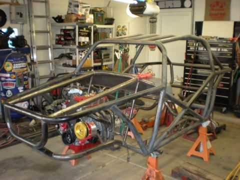Suzuki Samurai - A bare tubular steel roll cage/chassis for a custom vehicle build sits on orange jack stands in a garage, with a red and gold engine mounte…