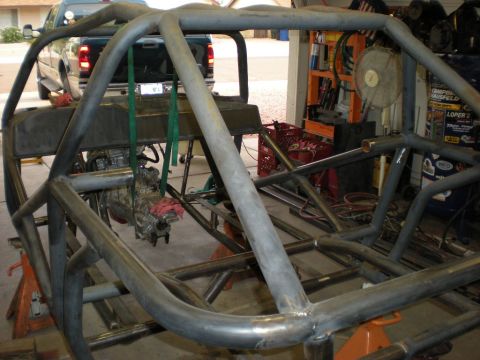 Suzuki Samurai - A bare tube chassis and full roll cage under construction on jack stands in a home garage, welds still raw and unpainted. A transmission an…