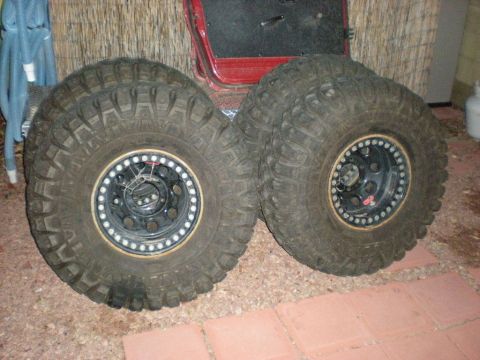 Suzuki Samurai - Two aggressive-tread mud/crawler tires mounted on black steel beadlock wheels with bolted gold-anodized lock rings, leaning against a reed…