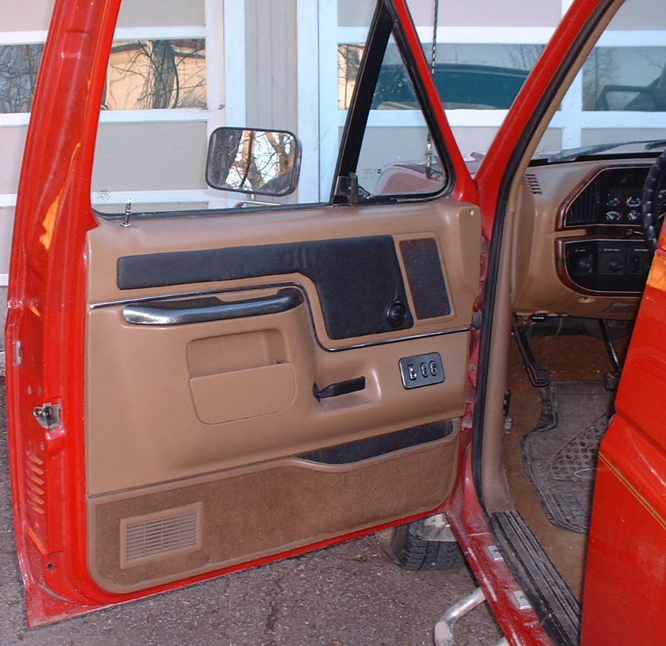 Interior view of a vehicle's driver side door panel and dashboard, showing tan and black trim, door handle, and window controls