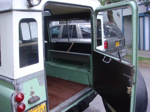 1967 Land Rover Land Rover - The rear of a 1967 Land Rover Series IIA is shown with its doors open, revealing the interior and two other vehicles parked behind it.