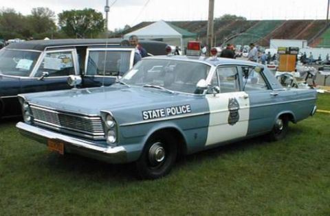 1991 Ford Crown Victoria - A vintage police car is parked on grass next to a hearse, with a stadium in the background.