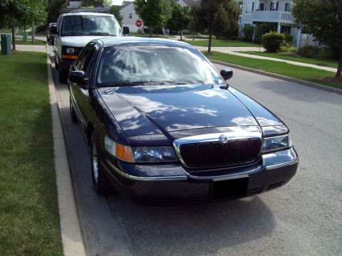 1999 Mercury Grand Marquis - A dark blue sedan is parked on the street next to a curb, with a white van behind it.