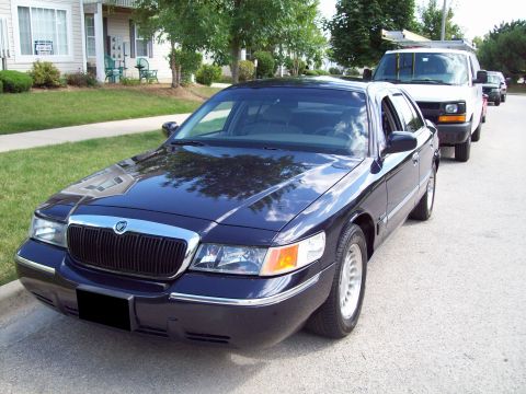 1999 Mercury Grand Marquis - A dark blue 1999 Mercury Grand Marquis is parked on a residential street, with a white van and another vehicle behind it.