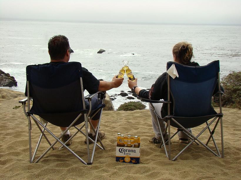 Two people in camping chairs on sand, toasting Corona beer bottles with ocean in background