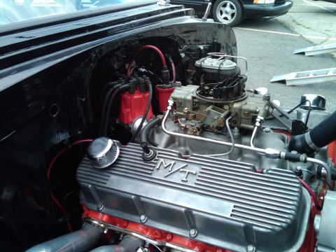 1955 Chevrolet Bel Air - Close-up of a V8 engine with M/T valve covers, showing the carburetor and distributor. Another vehicle is partially visible in the backgrou…