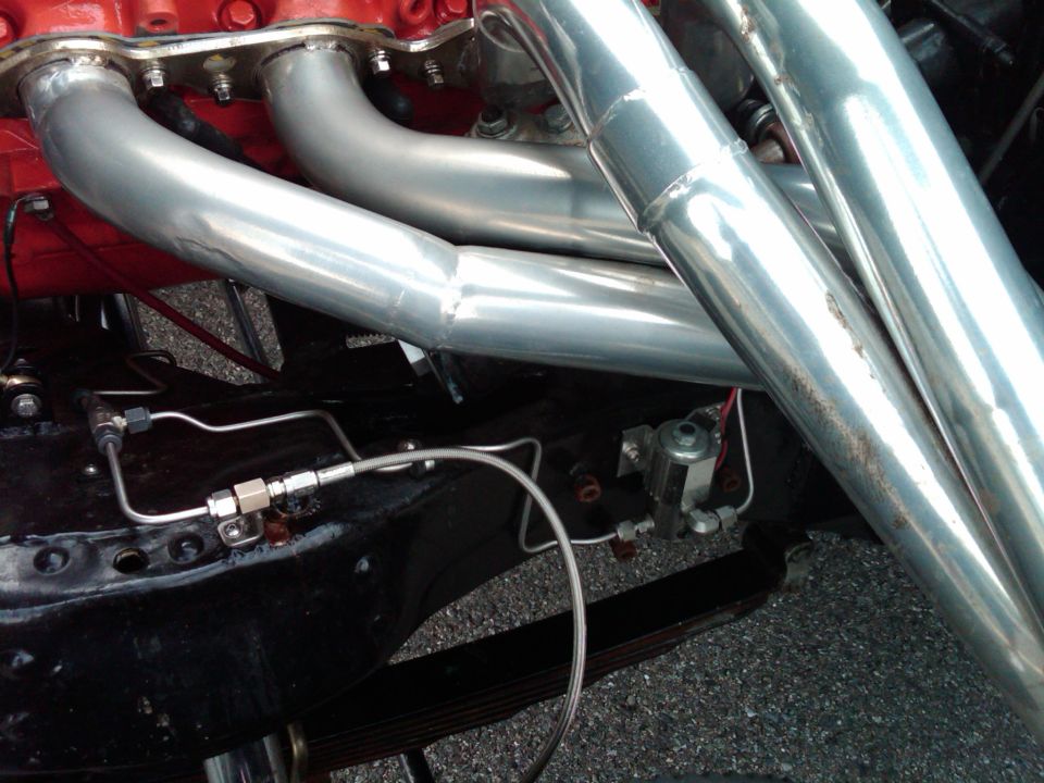 Polished long-tube headers on a red V8 with brake lines along the black frame rail