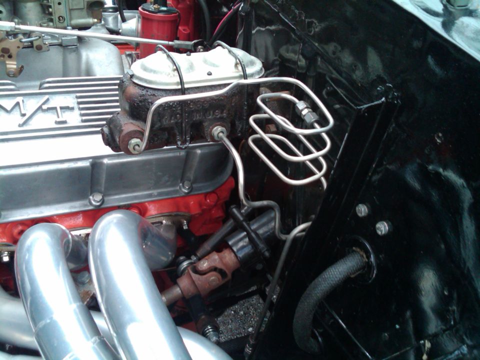 Close-up of a car's engine bay, showing the brake master cylinder, valve covers, and exhaust headers