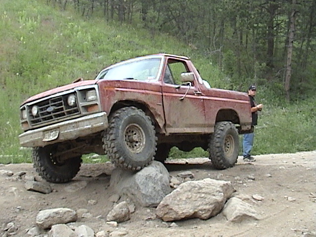 Red topless 1980s Ford Bronco on big tires climbing a boulder on a forest trail