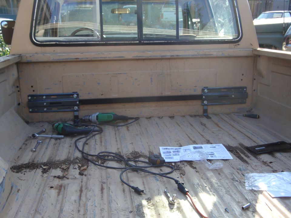 Tan pickup bed with black mounting brackets being installed on front bulkhead; drill and tools on bed floor