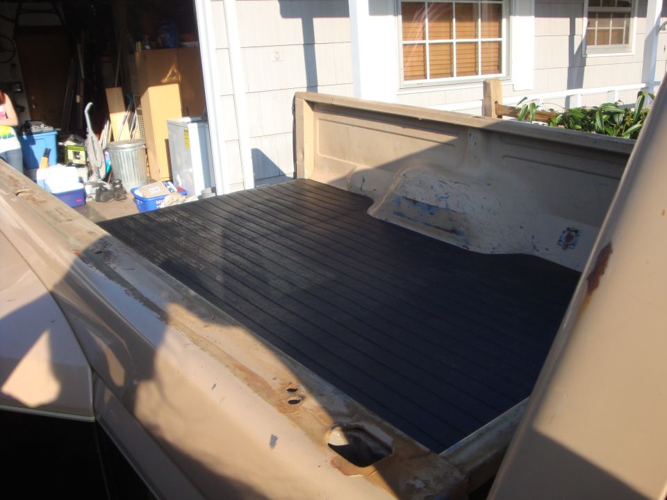 Truck bed with black ribbed liner, showing some rust on the bed rails. Garage and house in background