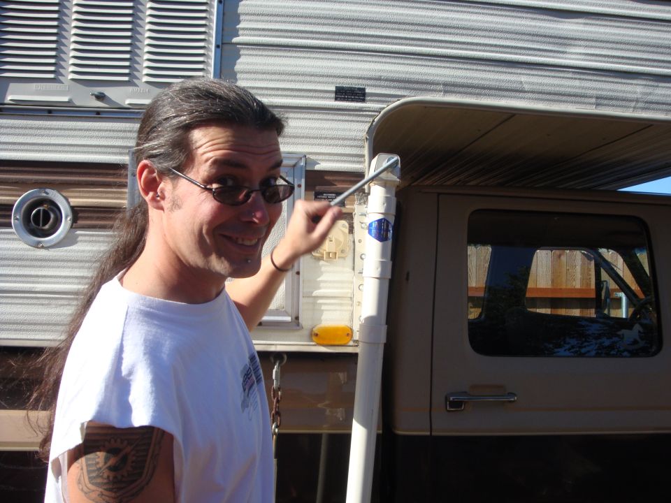 Man with long hair and sunglasses points to a white pipe next to a camper and a truck