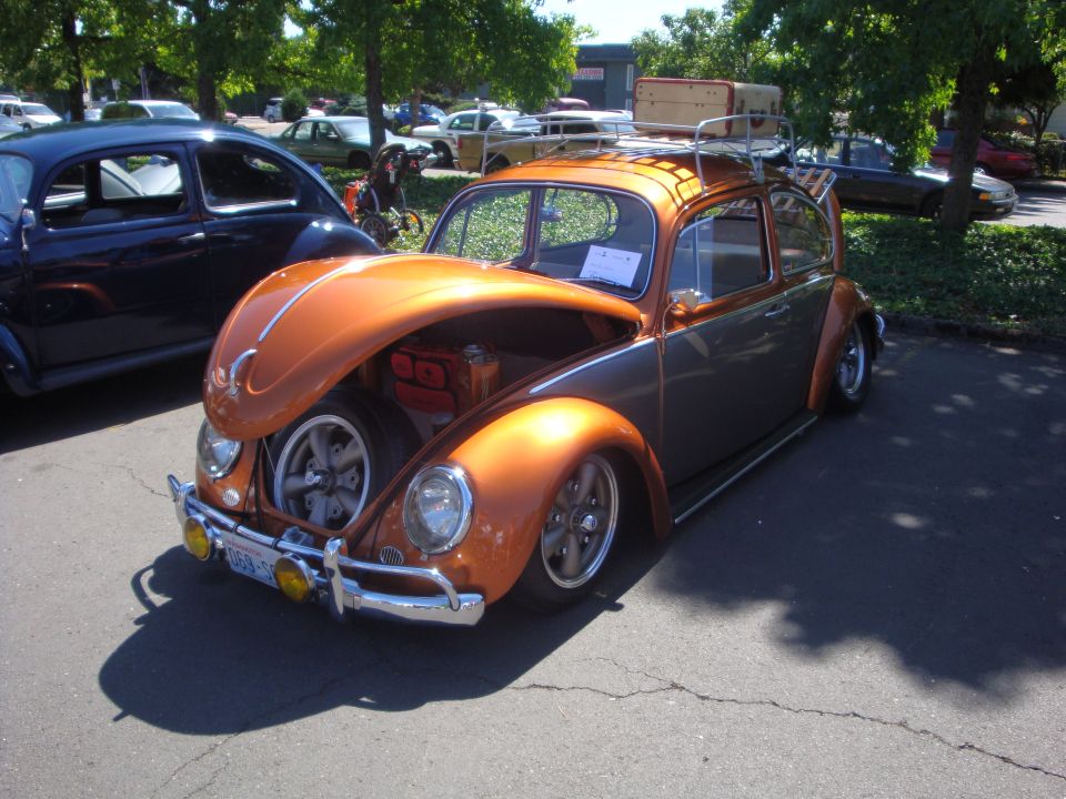 Orange and grey Volkswagen Beetle with luggage rack and suitcase on top. Front hood is open showing spare tire