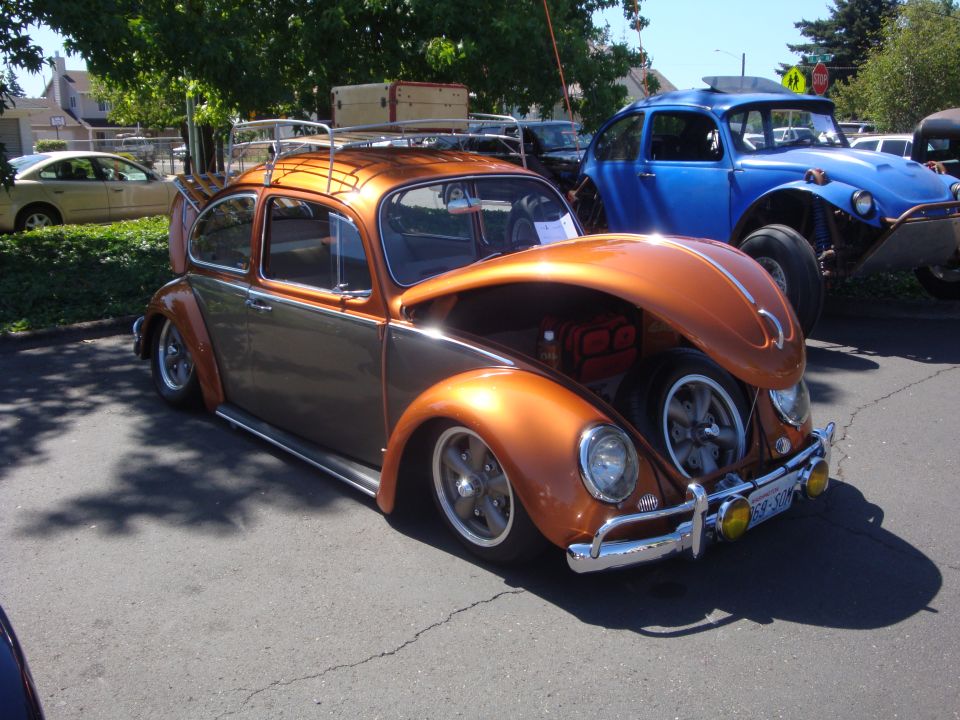 Orange and gray VW Beetle with hood open, luggage rack on top, parked next to a blue dune buggy