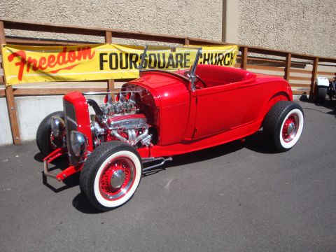 1964 Ford Fairlane - A bright red hot rod roadster is parked in front of a church with a banner.