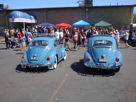 1964 Ford Fairlane - Two blue Volkswagen Beetles are parked at an outdoor event with people milling around tents.