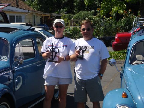 1964 Ford Fairlane - Two people with trophies pose with classic Volkswagen Beetles at an outdoor event.