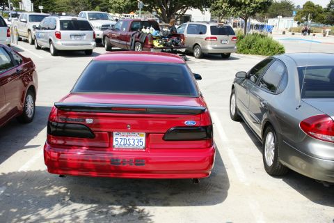 1990 Ford Taurus - A red Ford sedan is parked in a lot, showing its rear end with a spoiler and custom taillights.