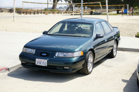 1990 Ford Taurus - This dark green sedan, likely a 1996 Ford Taurus, is parked near a beach volleyball court.